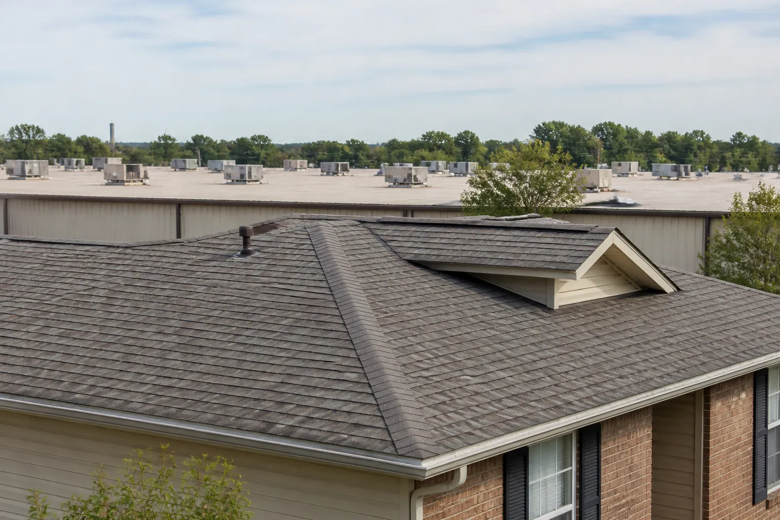 Asphalt-shingle roof with visible ridge, eaves, and flashing in the foreground and a low-slope commercial roofline in the distance.